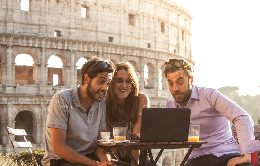 Three happy friends having video call sitting at table in front of colosseum in rome.