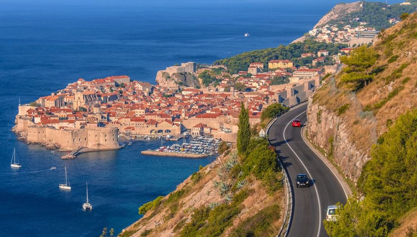 Photo of a road on the side of a mountain, overlooking the ocean, with a village in the distance