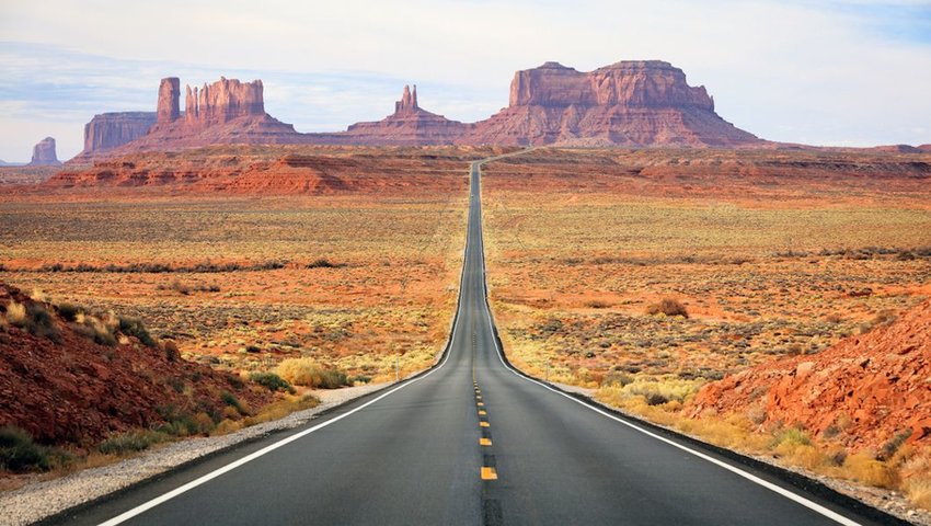 Photo of a road leading off into the distance, with red rocks towering in the sky