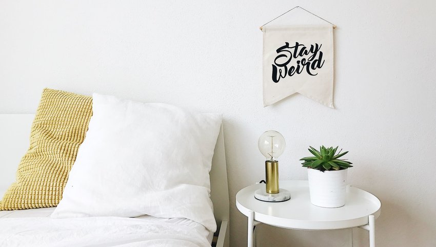 White bed with pillows next to a nightstand and a sign that says "Stay Weird"