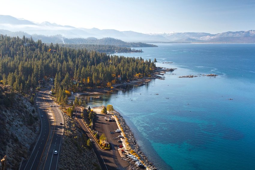 A road winding through trees with blue lake to the right and mountain range in background