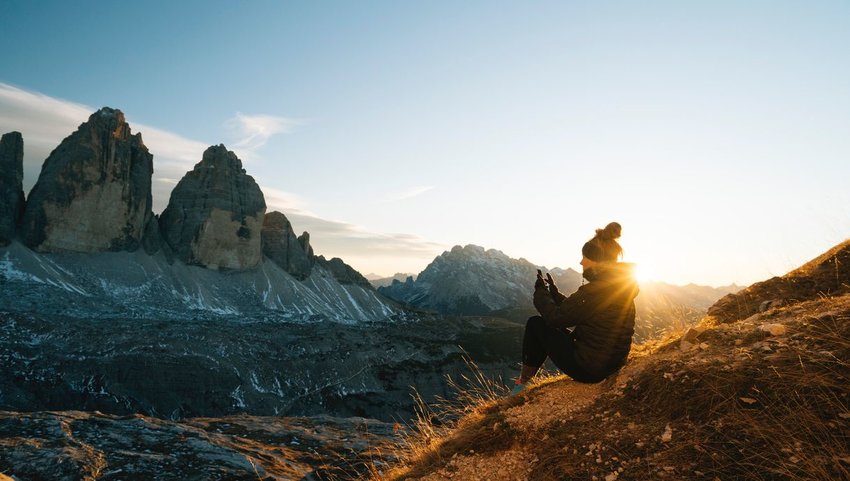 Person sitting on hill taking a photo on their phone of mountains