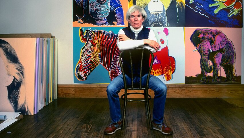 Artist Andy Warhol seated in front of some of his artwork.