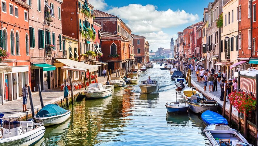 Canal lined with boats and people walking on sidewalks. 