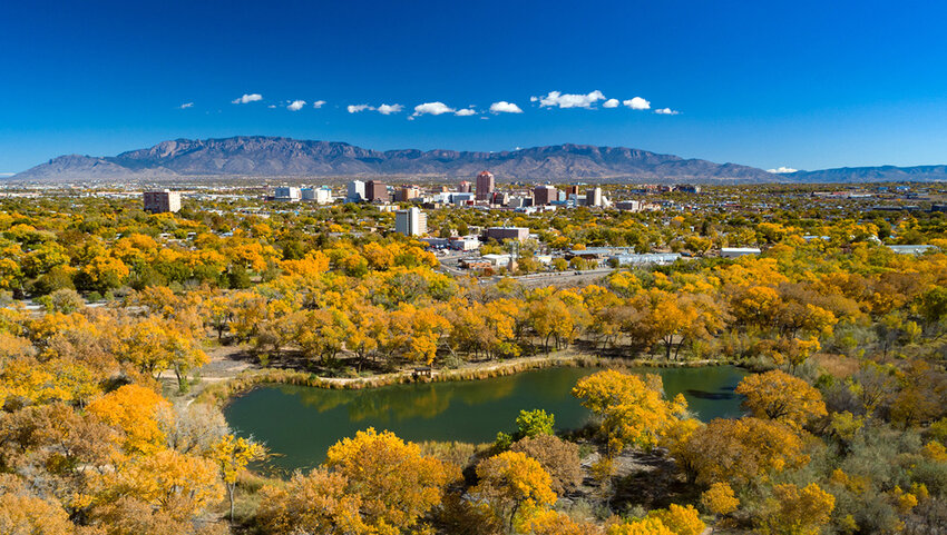 Albuquerque during autumn with the downtown skyline and the Sandia Mountains in the distance, with golden trees and a small lake in the foreground.