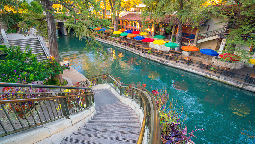 Stairs down to river walk, with river and row of tables and colorful umbrellas.