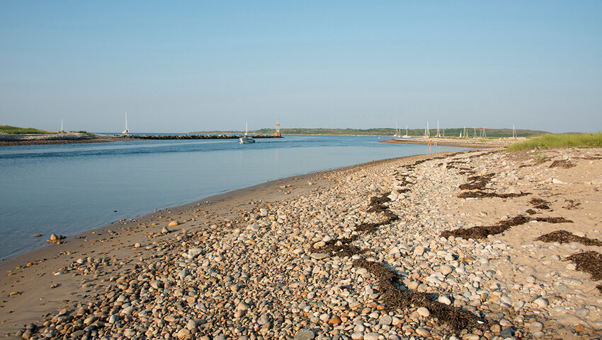 Rocky shoreline with boats in harbor in distance. 